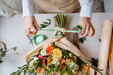 A Female Florist Ties A Green Ribbon Bow On A Bouquet Of Flowers Wrapped In Craft Paper On The Desktop. Top View.