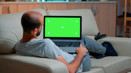 Poster - Tired man lying on sofa working on computer with mock up green screen chroma key display searching online information. Caucasian male using isolated pc late at night in kitchen