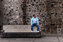 Young Mexican Bearded Man Wearing A Light Blue Dress Shirt Sitting In A Bench Outside A Church With A Huge Stone Wall In Cholula, Mexico. Wide Shot