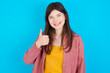 young beautiful Caucasian woman wearing pink T-shirt over blue wall giving thumb up gesture, good Job! Positive human emotion facial expression body language.