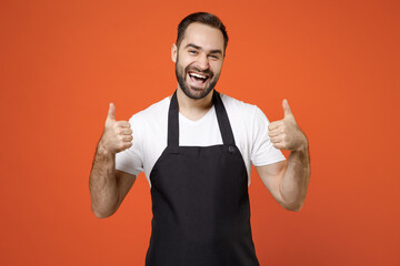 Young happy man barista bartender barman employee in black apron white t-shirt work in coffee shop sshow thumb up gesture isolated on orange background studio portrait Small business startup concept.