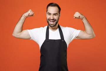 Wall Mural - Young man barista bartender barman employee in black apron white t-shirt work in coffee shop show biceps hand muscles demonstrating strength power isolated on orange background Small business startup.