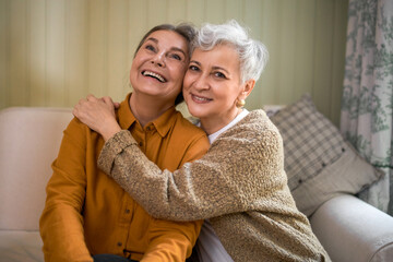 Friendship, family and relationships concept. Happy carefree mature sisters bonding, sitting on comfortable couch at home, hugging, expressing love and care. Two elderly best friends having fun