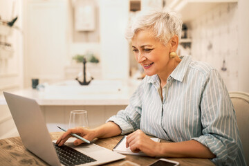 Attractive middle aged female freelancer in striped shirt sitting at home in front of open laptop, typing, working on content for website, using high speed internet connection, enjoying online work