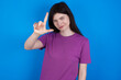 © Roquillo - young beautiful Caucasian woman wearing purple T-shirt over blue wall making fun of people with fingers on forehead doing loser gesture mocking and insulting.