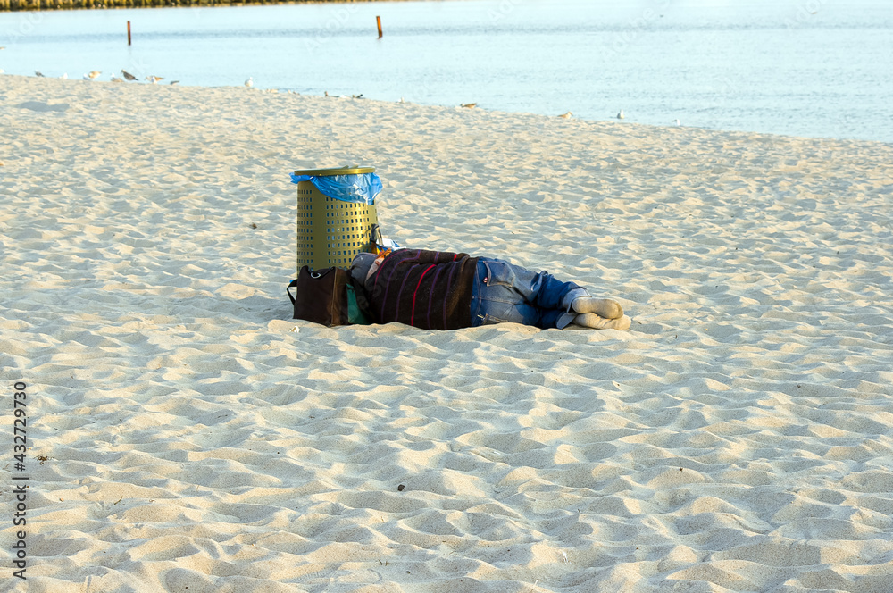 Samotny bezdomny człowiek śpiący na piaszczystej plaży w pobliżu pojemnika na śmieci - obrazy, fototapety, plakaty 