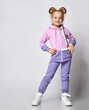 Studio shot of a small blonde girl in pink violet tracksuit with a hood, posing for the camera with her hand, staring at the camera, isolated on white background.