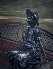 Black Metal Sculpture. Girl In Vintage Dress Sitting On A Park Bench Side View On Green Blurred Background Closeup. Selective Focus