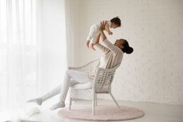 Wall Mural - overjoyed young african american mother sit in chair at home play with small toddler baby daughter. 