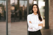 © diignat - Portrait of a Caucasian pretty business lady smiling to the camera and crossing her hands while standing on the glass building background outdors