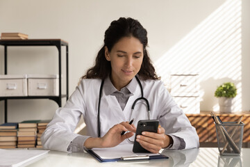 Wall Mural - Consulting patient by internet. Focused female medic sit at workplace hold phone text message to client check health status on distance. Woman doctor work with smartphone read email from sick person