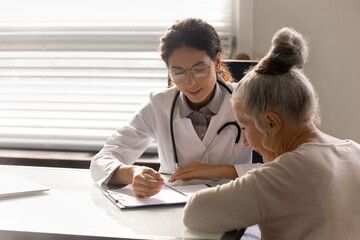 Wall Mural - Consulting patient. Capable young latin woman doc counsel female retiree prescribe treatment explain therapy details. Old lady sign informed consent on surgical procedure at attending physician office