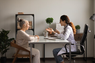 Wall Mural - Medicare for older people. Friendly hispanic woman gp family therapist consult elderly female patient at meeting at hospital cabinet. Old lady retiree visit doc listen to recommendation for treatment