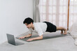 © Gengwit - Training At Home. Sporty man doing yoga while watching online tutorial on laptop, exercising in living room, free space