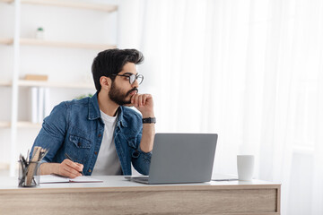 Thoughtful arab freelancer noting ideas to notepad, taking notes, sitting at workplace with laptop computer, copy space