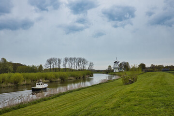 Wall Mural - Horizontal view on small river Linge with a white boat and a meadow with a white Dutch mill on a cloudy day. Betuwe in the Netherlands with copy space.