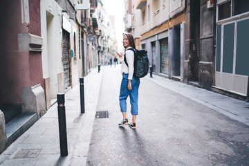 Wall Mural - Full length portrait of cheerful woman walking at city street and smiling at camera, happy female tourist with smartphone gadget using roaming internet for searching location of quarter in town