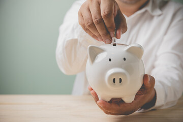 Asian adult businessman holding the white piggy bank on hands, and putting coin into piggy bank on a wooden table. Concept financial, accounting, savings the money, and loan for investment business.