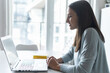 © Vadim Pastuh - Smiling woman using laptop at home, she sits at the desk with a panoramic window on the background. Female employee, freelancer working remotely from home, has video meeting, video call