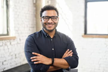 Headshot of skilled hindu male employee standing with arms crossed in modern office, successful confident mixed-race man wearing eyeglasses and smart casual, business portrait of indian entrepreneur