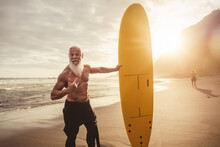 Senior Man Having Fun Surfing During Sunset Time - Retired Male Training With Surfboard On The Beach - Elderly Healthy People Lifestyle And Extreme Sport Concept