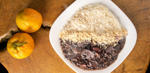 Brazilian feijoada, white dish with a delicious Brazilian feijoada, rice and farofa, with two oranges on rustic wood, top view.