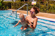 © Alyona - Tanned smiling woman in swimsuit teaches to swim one-year-old baby girl in outdoor pool