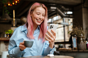 Wall Mural - Young smiling woman using mobile phone while drinking coffee in cafe