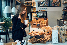 Woman holding cup with coffee looking at pastry, buns, cakes and cookies and waiting for the order. Girl buying a sweet food and hot drink to go. Young woman having a break