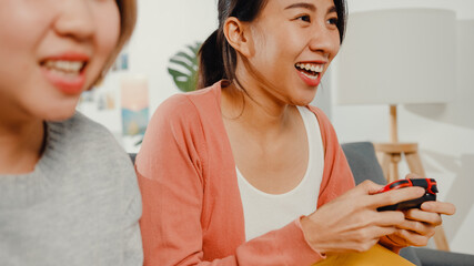 Wall Mural - Lesbian LGBTQ women couple play video game at home. Young Asia lady using wireless controller having funny happy moment together on sofa in living room. They have great and fun time celebrate holiday.