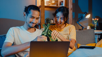 Sticker - Happy young asian couple enjoy night party event online sit couch use laptop computer video call with friends toast drink beer via video call online in living room at home, Social distancing concept.