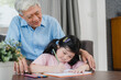 © tirachard - Asian grandfather teach granddaughter drawing and doing homework at home. Senior Chinese, grandpa happy relax with young girl lying on sofa in living room at home concept.