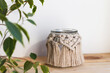 Beautiful jar candle holder with macrame decor on wood shelf. Boho home decor. Houseplant in front. Rustic decoration.