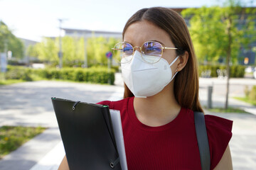 Wall Mural - Female university student with FFP2 KN95 face mask walking in city street. College girl back to school holding bag and folders.