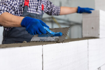 The master performs construction work, namely, laying a wall of gas blocks with tools and adhesive mortar, in the background the sky and the forest.