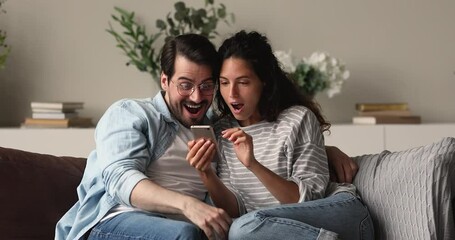 Wall Mural - Sincere affectionate young family couple reading email or mobile notification, feeling excited of getting amazing news, celebrating online lottery win together, relaxing on comfortable sofa at home.