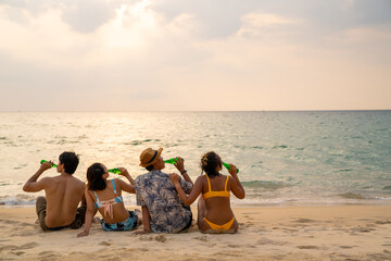 Wall Mural - Group of Happy Asian man and woman friends sitting on the beach enjoy drinking beer with talking together at summer sunset. Male and female friendship relax and having fun outdoor lifestyle activity.