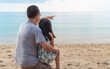 Asian father and little daughter together on the beach, dad teaching by pointing to the horizon, child sitting on father's lap, back side of father and child, blank space for copy and design.