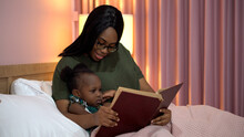 African American Mother Reading A Book To Her Little Daughter In Bed Before Going To Sleep At Home. Black Mom Story Telling To Girl In Bedroom