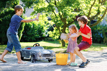 Wall Mural - Three happy children washing big old toy car in summer garden, outdoors. Two boys and little toddler girl cleaning car with soap and water, having fun with splashing and playing with sponge.