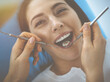 © rogerphoto - Smiling brunette woman being examined by dentist at dental clinic. Hands of a doctor holding dental instruments near patient's mouth. Healthy teeth and medicine concept