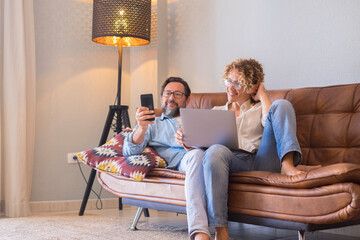 Wall Mural - People at home with computer and smart phone enjoy time together - happy couple young adult laying and sitting on the sofa closer - life and relationship man and woman with internet wireless