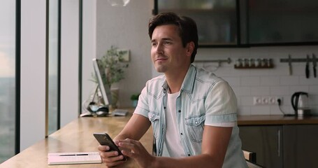 Young man sit in kitchen using smartphone, learn new mobile application, web surfing, check personal organizer, plan meeting, send e-mail solve business remotely, think over response to client concept