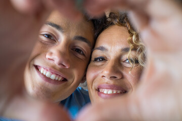 Wall Mural - Portrait of couple of mom and son together a home making a heart love shape with their fingers and hands with the faces at the background. Affectionate teenager and woman enjoying
