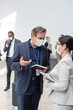 © LIGHTFIELD STUDIOS - interracial business people in medical masks, with notebook and digital tablet, talking in hall during conference