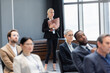 © LIGHTFIELD STUDIOS - Businesswoman with paper folder and cellphone standing near multiethnic colleagues on blurred foreground