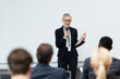 © LIGHTFIELD STUDIOS - Mature speaker with paper folder talking near blurred business people during meeting