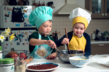 Children, dressed in aprons and cooking hats in a cosy kitchen are mixing daugh with spoons in a bowl with different kitchen implements on the table. Cooking kids