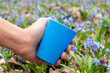 © Kathrine Andi - Hand holding blue reusable plastic glass for picnic in wild spring blooming lawn close-up. Travel cup in blue flowers with focus blur. Eco picnic utensil details.