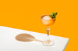 Elegant stemware glass of fresh orange cocktail with big ice ball on white table surface, bright yellow background
